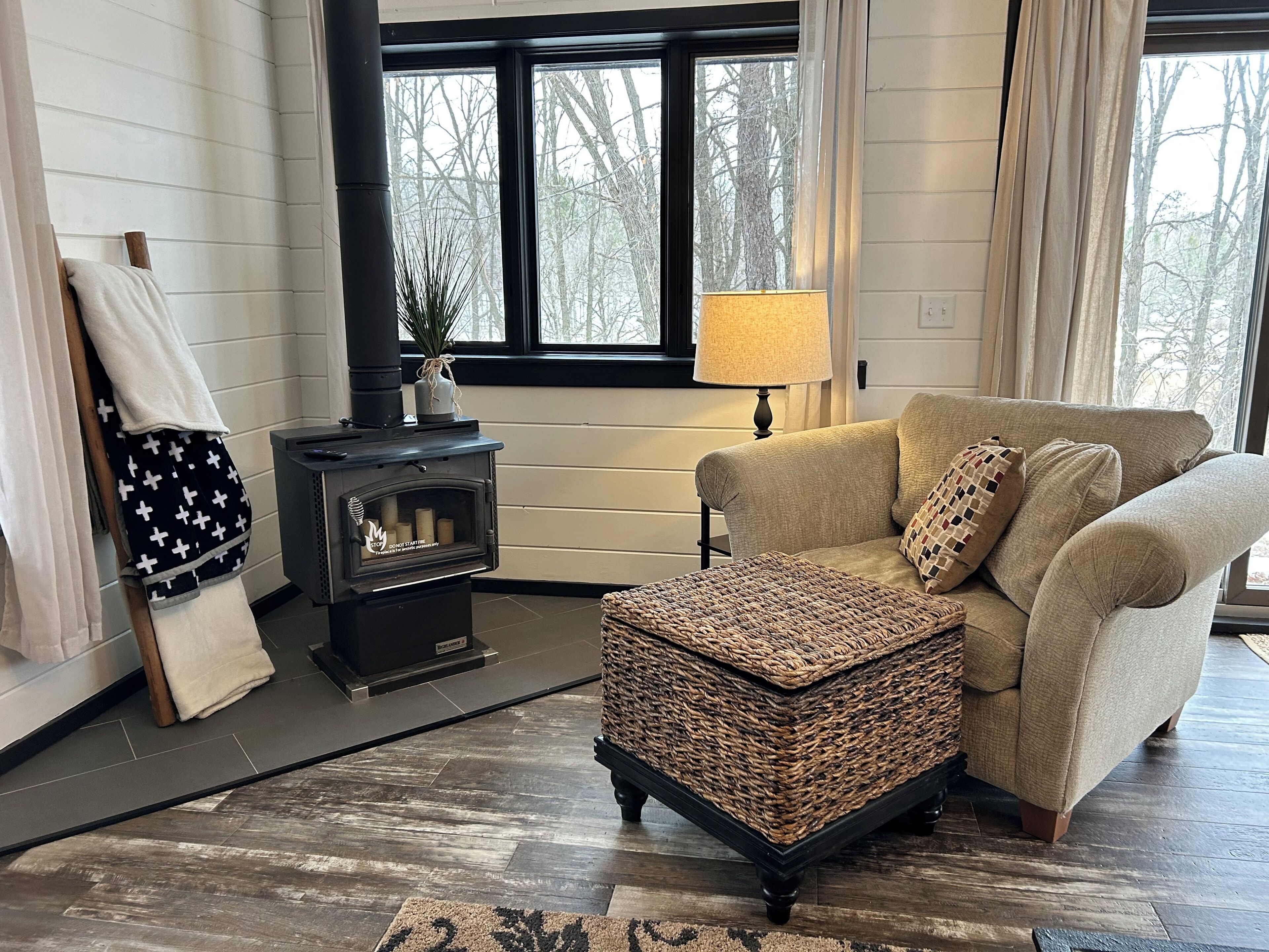 A cozy living room with a sofa, a woven ottoman, a fireplace, and a lamp.