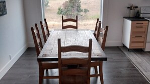Un comedor con una gran mesa y sillas de madera, una cocina con mostrador y armarios, y vistas a un paisaje rural a través de un ventanal.
