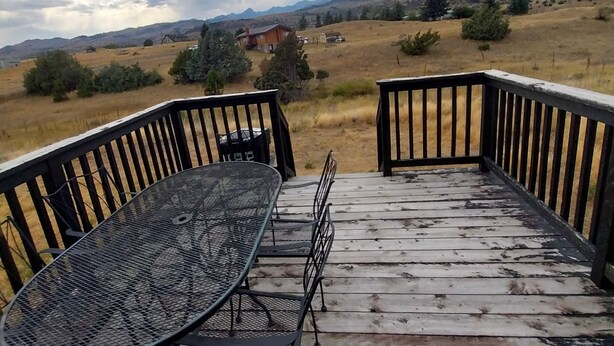 Una terraza de madera con una mesa y sillas de metal, con vista a un paisaje cubierto de césped, colinas lejanas y una casa.