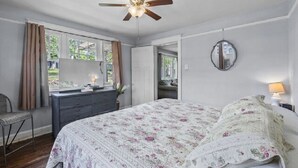 A bedroom with a bed, a dresser, a chair, a ceiling fan, a window with curtains, and a mirror on the wall. - AV    Bungalows   #7 (Arcadia)