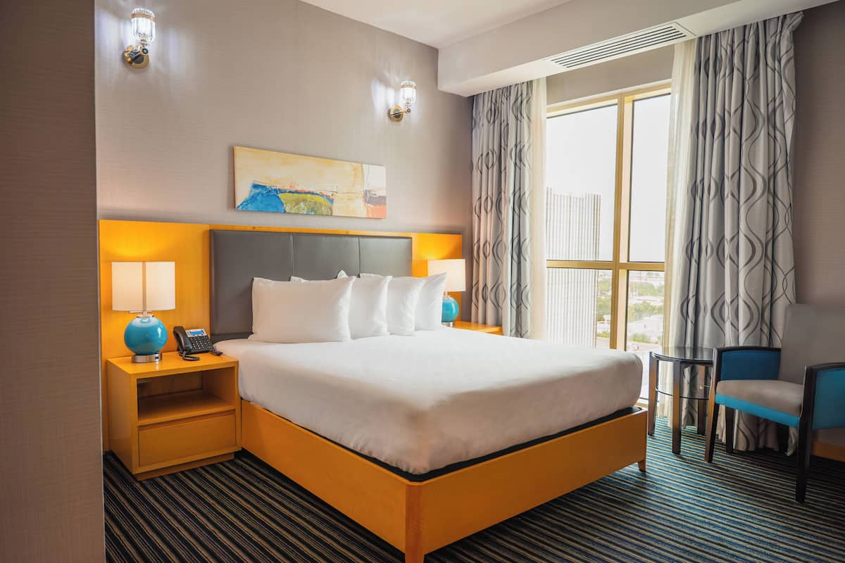 Habitación de hotel con una cama grande, dos mesitas de noche con lámparas, una silla, un cuadro en la pared y una ventana con cortinas.