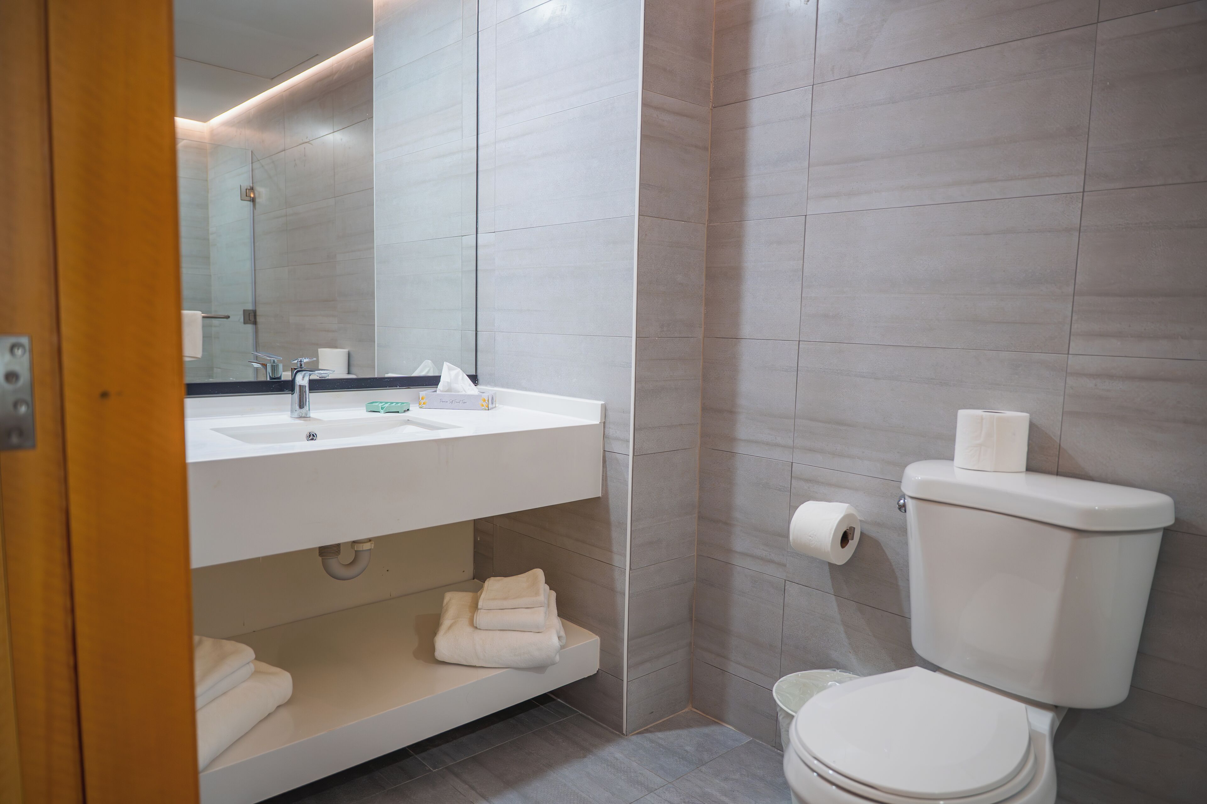 A modern bathroom with a white sink, mirror, and toilet. There is a towel rack and a shelf with folded towels.