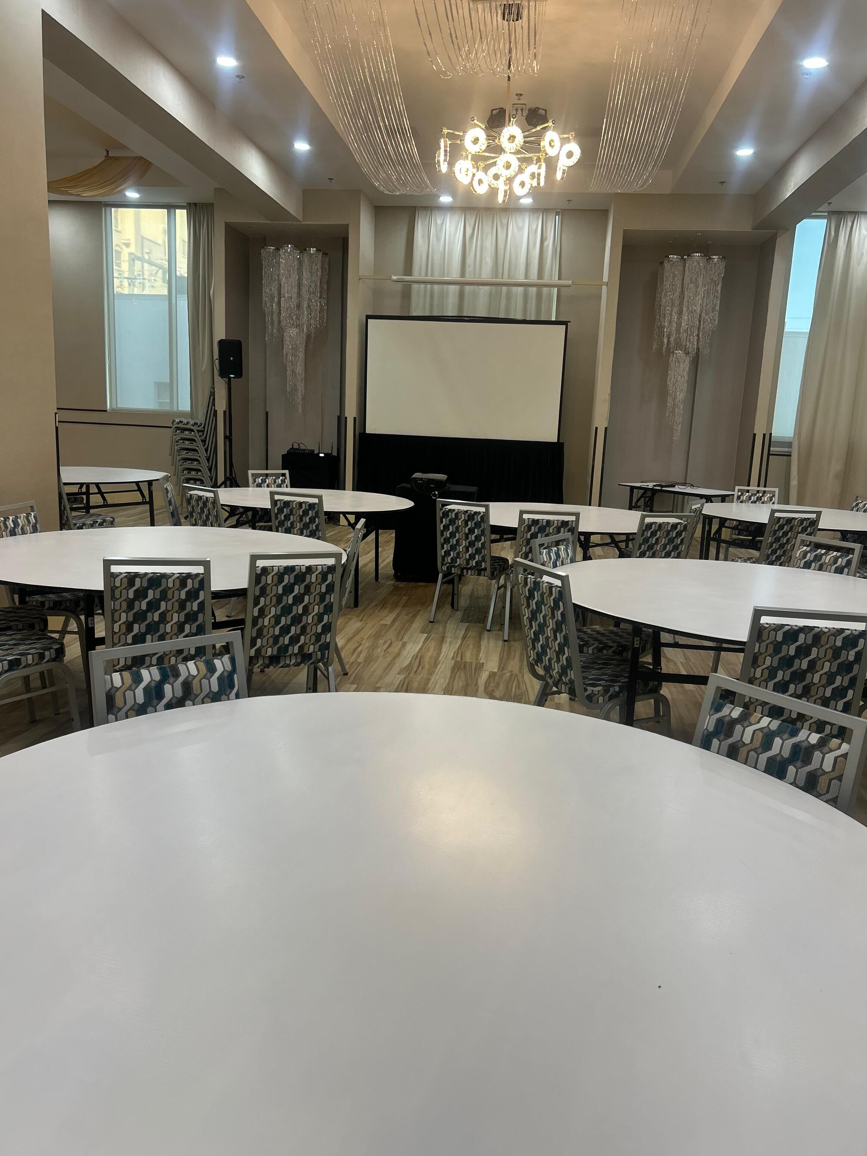 A conference room with multiple tables and chairs, a projector screen, and a chandelier.