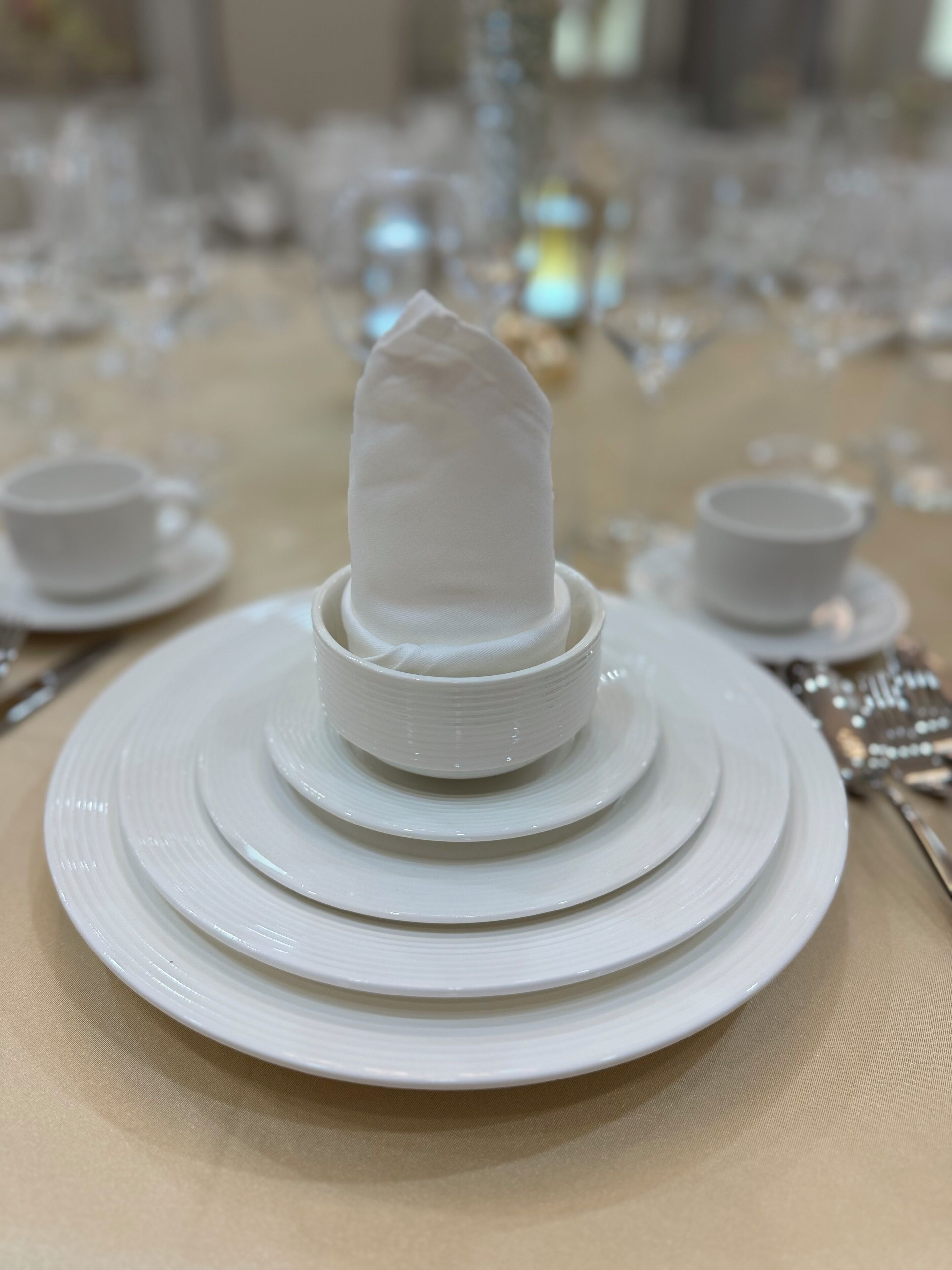 Uma mesa posta com pratos brancos, uma folha de servilleta branca no centro e uma xícara.