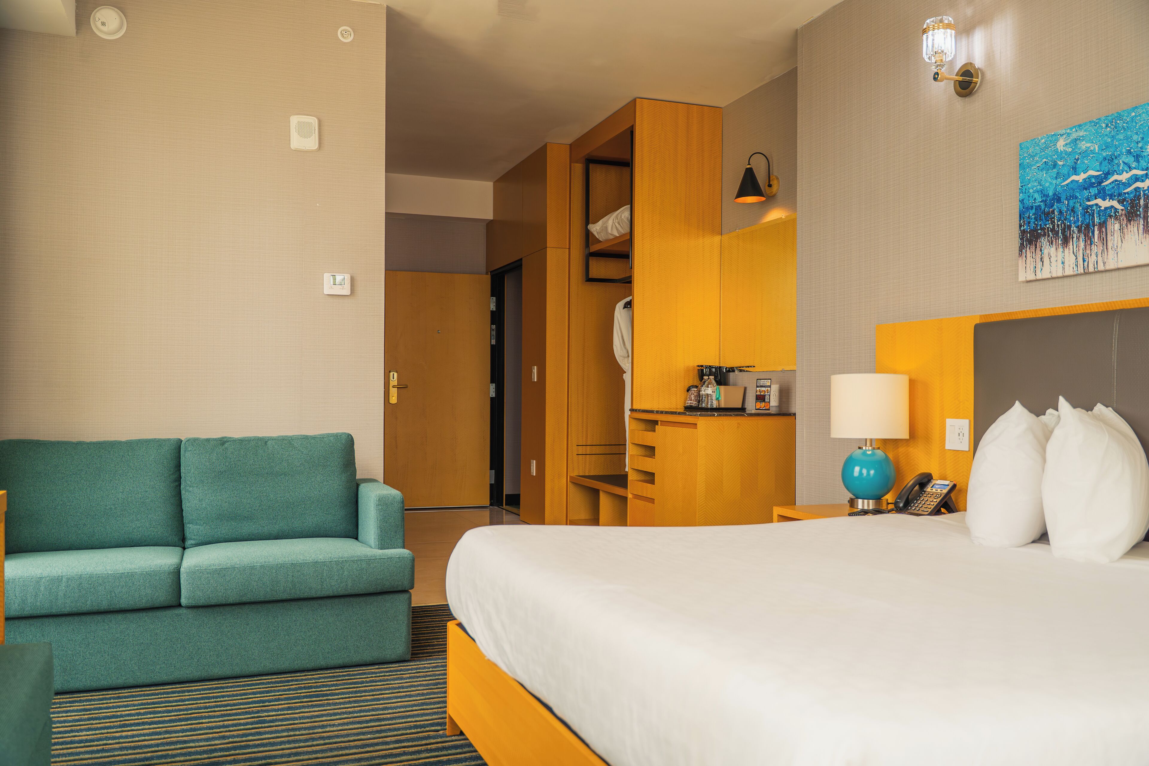 A hotel room with a bed, a green sofa, a desk with a lamp, and a painting on the wall.