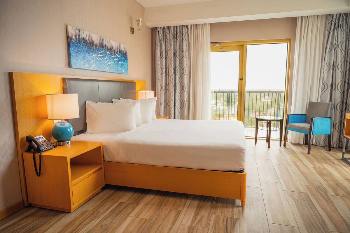 Habitación de hotel con una cama grande, dos mesitas de noche con lámparas, un escritorio pequeño con una silla y un balcón con cortinas.