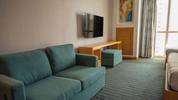 Et hotelværelse med en grøn sofa, en træbænk, et fladskærms-tv monteret på væggen og et stort vindue med mønstrede gardiner.
