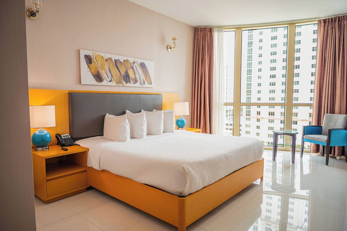 Habitación de hotel con una cama grande, dos mesitas de noche con lámparas, una silla y un ventanal con cortinas.
