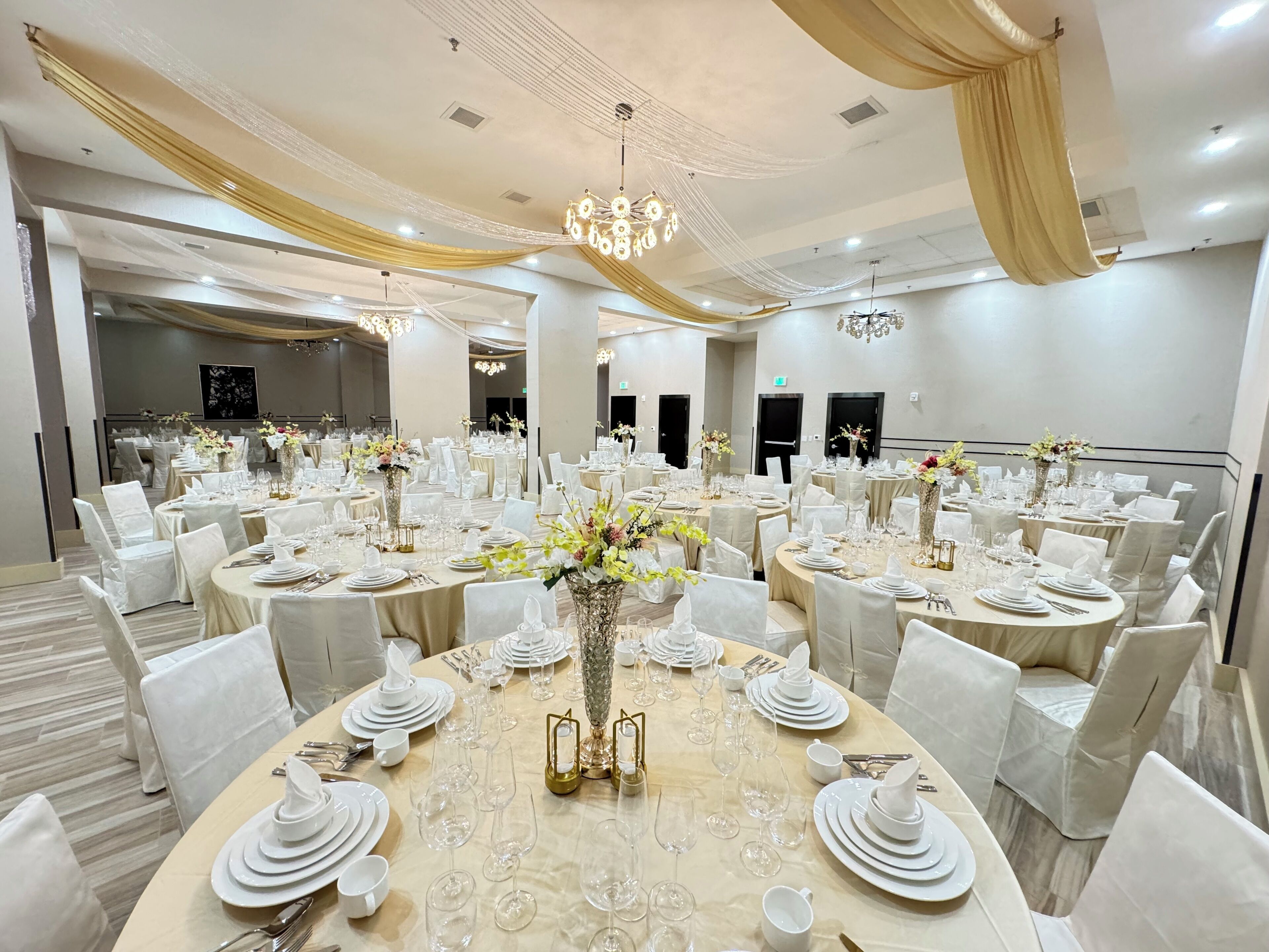 Une salle de banquet avec des tables rondes dressées pour un événement formel, agrémentée de nappes blanches, de tentures dorées et de lustres.