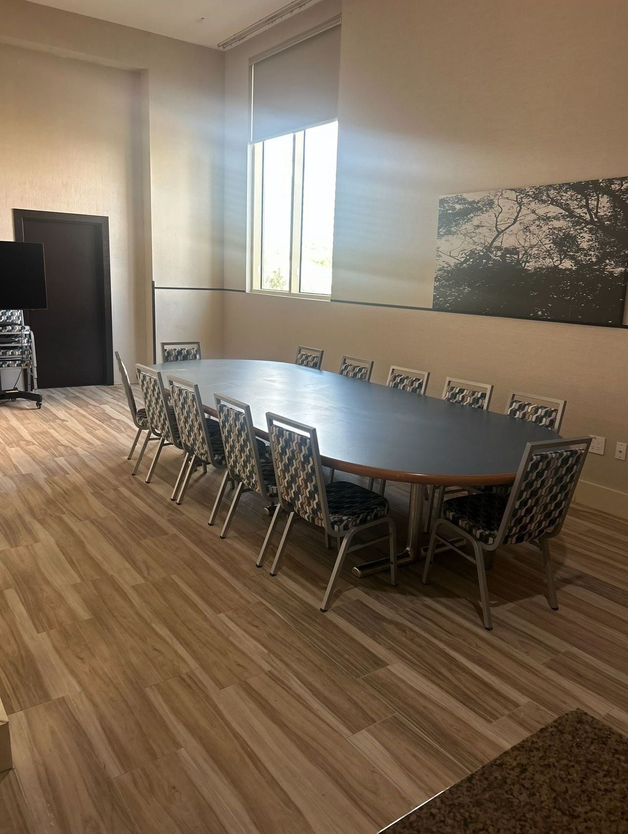 A conference room with a large oval table, patterned chairs, a large window, and a painting on the wall.