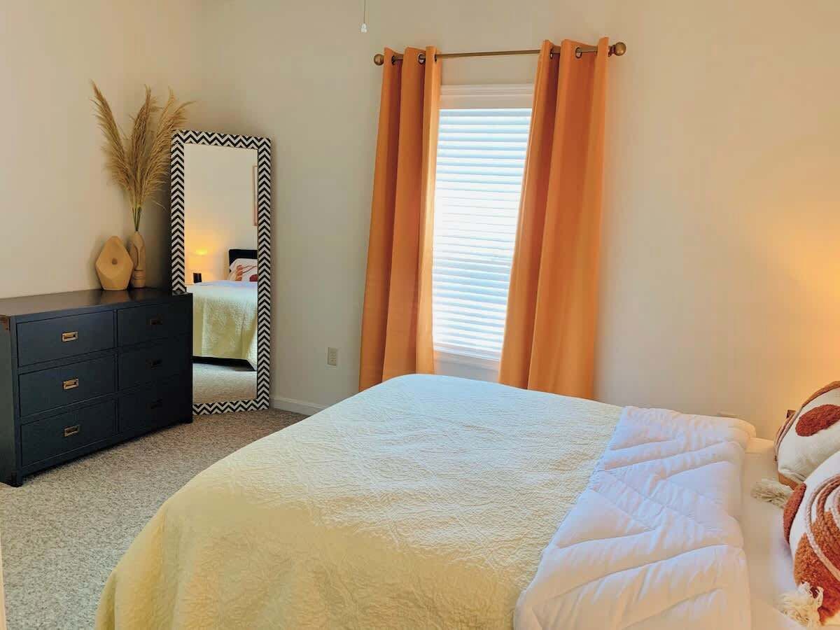A bedroom with a bed, a dresser, a mirror, and a window with curtains.