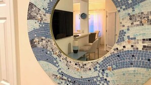 Et rundt spejl med mosaik-kant, der afspejler et rum med skrivebord, stol og et vindue med gardiner.