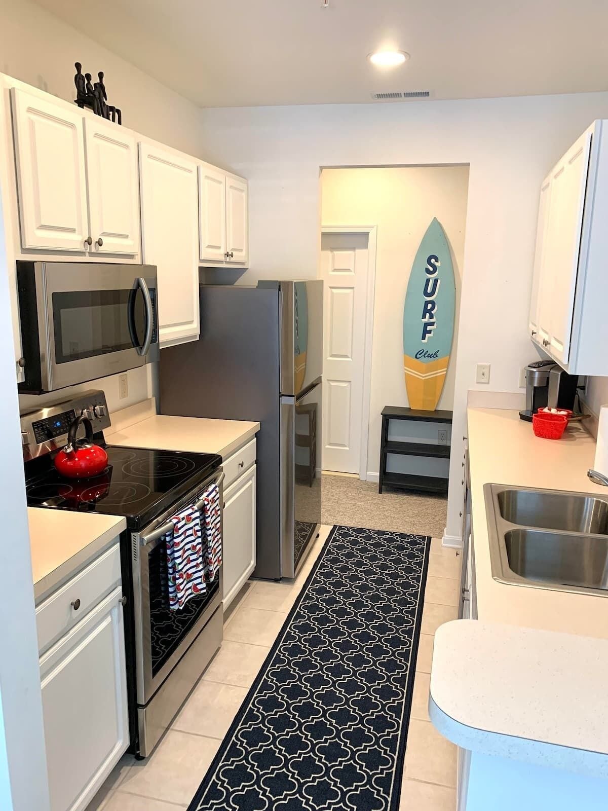 Una cocina moderna con armarios blancos, electrodomésticos de acero inoxidable, un hervidor rojo y un adorno con forma de tabla de surf.
