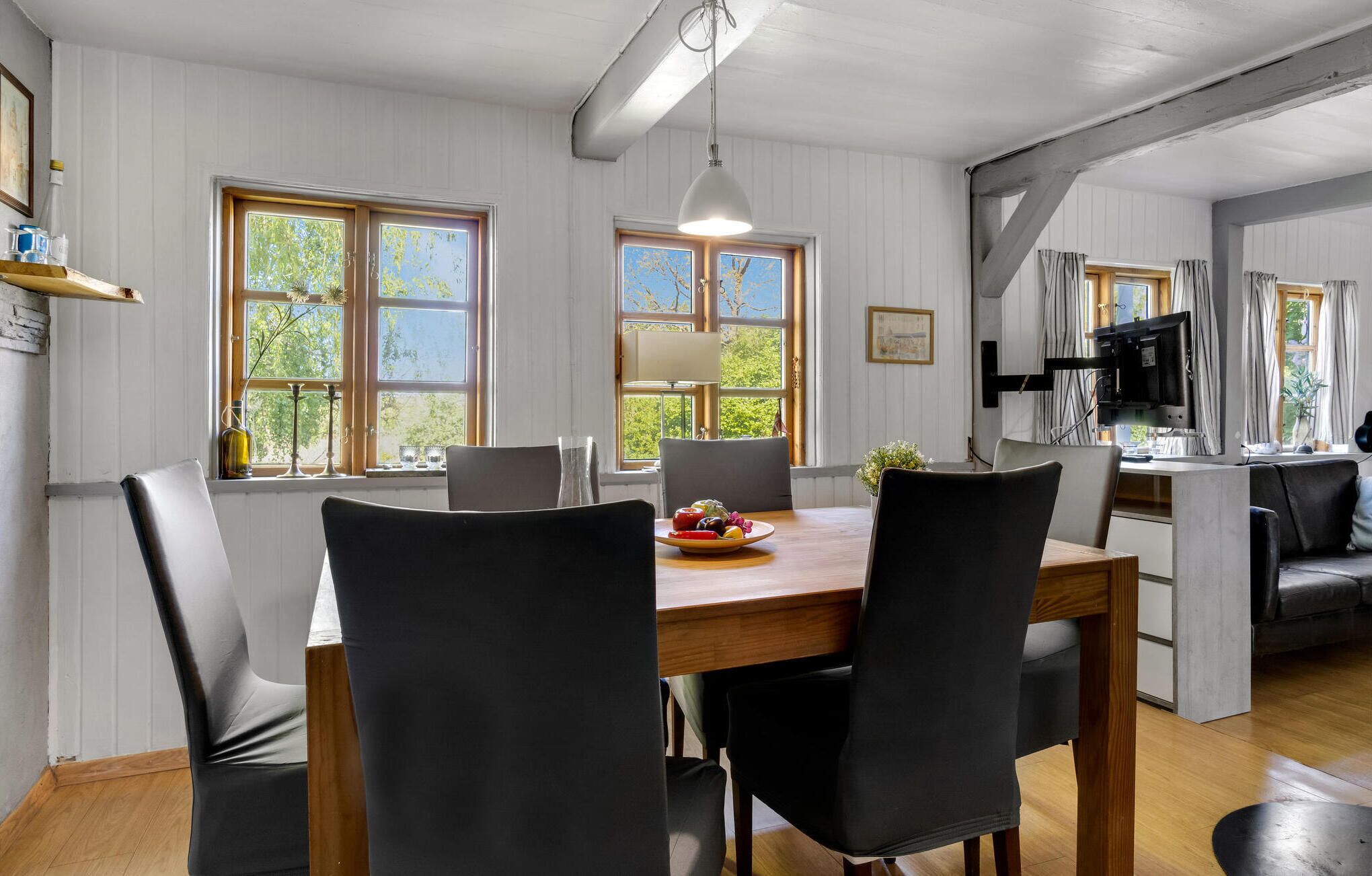 A dining area with a wooden table, black chairs, and a fruit bowl. There are windows with curtains, a television, and a shelf with items.