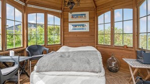 Ein Zimmer in einer Holzhütte mit einem Bett, einem Schreibtisch und zwei Stühlen. Große Fenster und zwei Bilder schmücken die Wand.