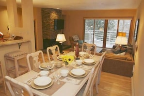 Un comedor con una mesa larga dispuesta para cuatro personas, rodeada de sillas blancas. Hay una pared de acento de piedra y un televisor montado sobre la chimenea.