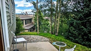 A patio with two white chairs and a small round table, surrounded by a concrete area and a grassy garden with trees.