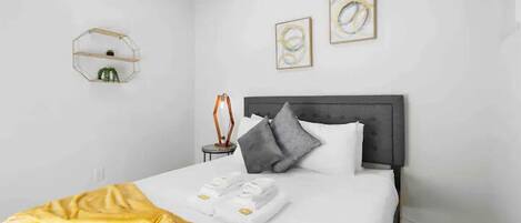 A neatly made bed with a gray headboard, a yellow throw blanket, and a pair of folded towels. There are two abstract paintings on the wall above the bed.
