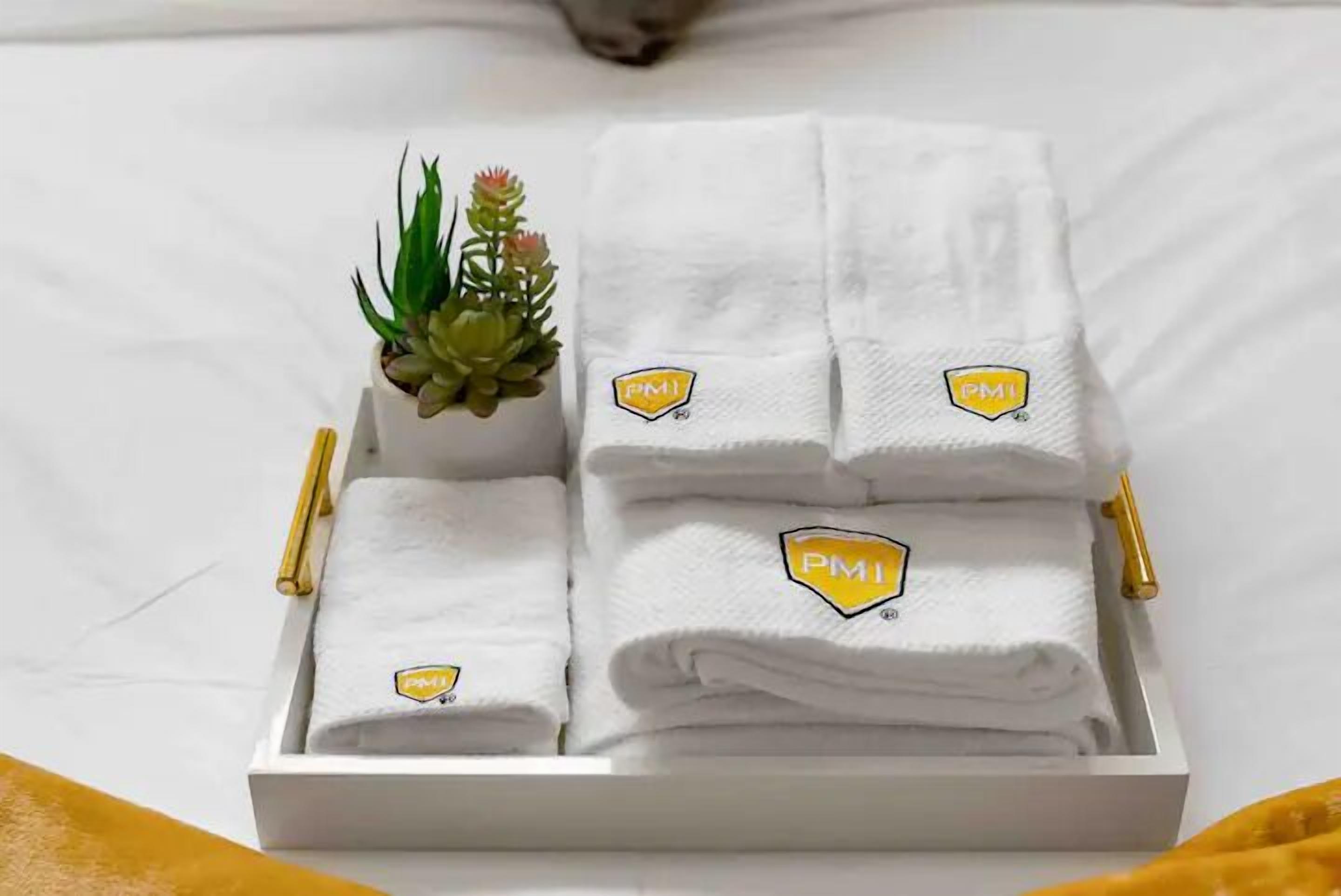 Uma bandeja branca com toalhas brancas dobradas, cada uma com o logotipo amarelo 'PMI', e uma pequena planta em vaso sobre uma superfície branca.