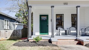 Une maison blanche avec une porte verte, portant le numéro 1012, et une véranda avec des chaises.
