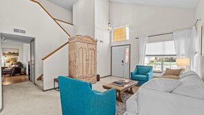 A spacious living room with a staircase, a large wooden cabinet, a blue armchair, a white sofa, and a window with curtains. - Location! Location! Amazing *Hidden Gem* -NE Heights! Family and pet friendly 🐶  (Albuquerque)