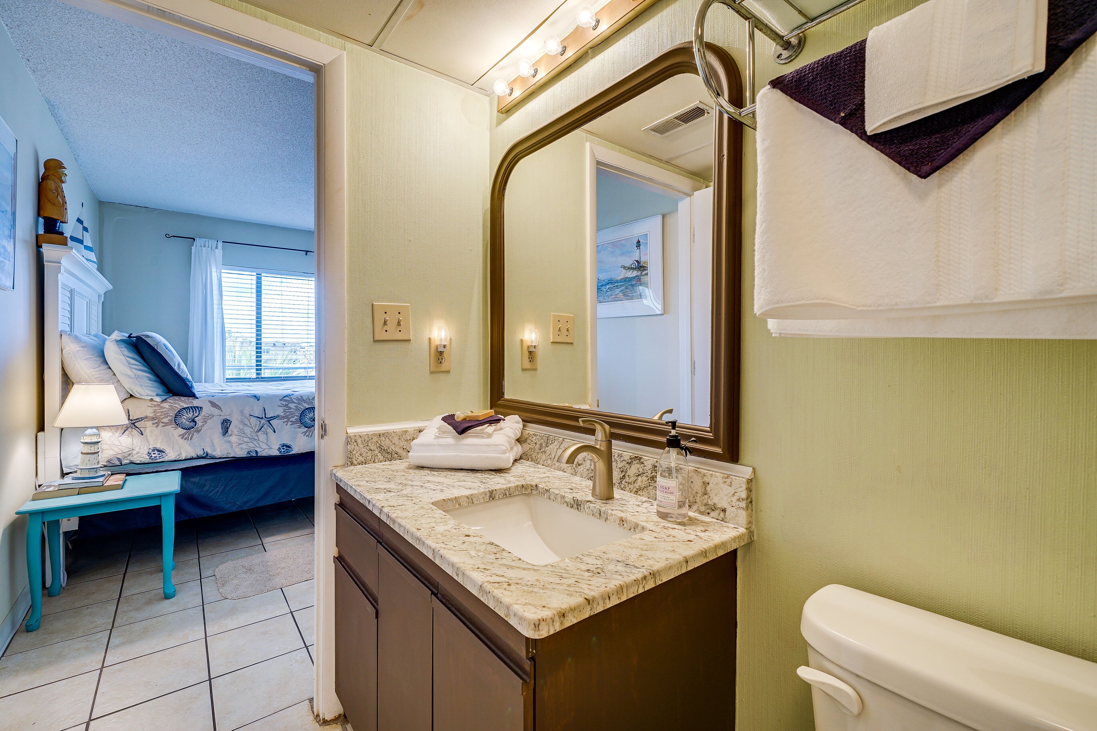 Een badkamer met een wastafel, spiegel en toilet. In de aangrenzende kamer staat een bed met een blauwe deken en kussens.
