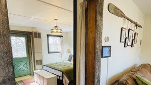 A room with a wooden ceiling, a bed with a green blanket, a bench, a brown leather couch, and a wall-mounted paddle. - Charming 1BR riverside cabin in Brainerd, MN. River sunsets, stocked kitchenette, fire pit, 10 min to downtown. Ideal for couples, small families & remote workers seeking a peaceful Up North escape. (Daggett Brook Township)