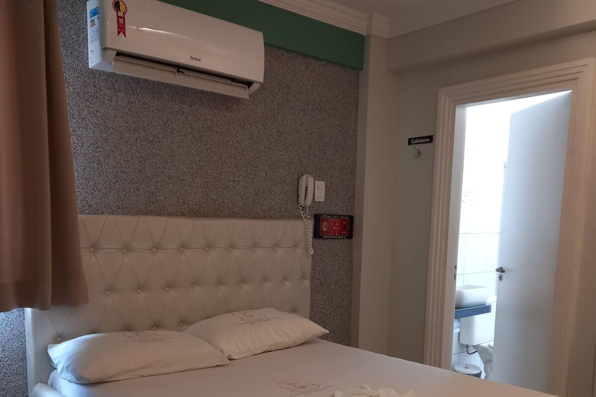 Habitación de hotel con cama, teléfono, equipo de aire acondicionado y puerta al baño.