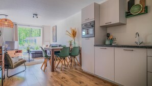 Eine moderne Küche mit weißen Schränken, einem eingebauten Ofen und einer Spüle. Es gibt einen Essbereich mit einem Holztisch und grünen Stühlen.