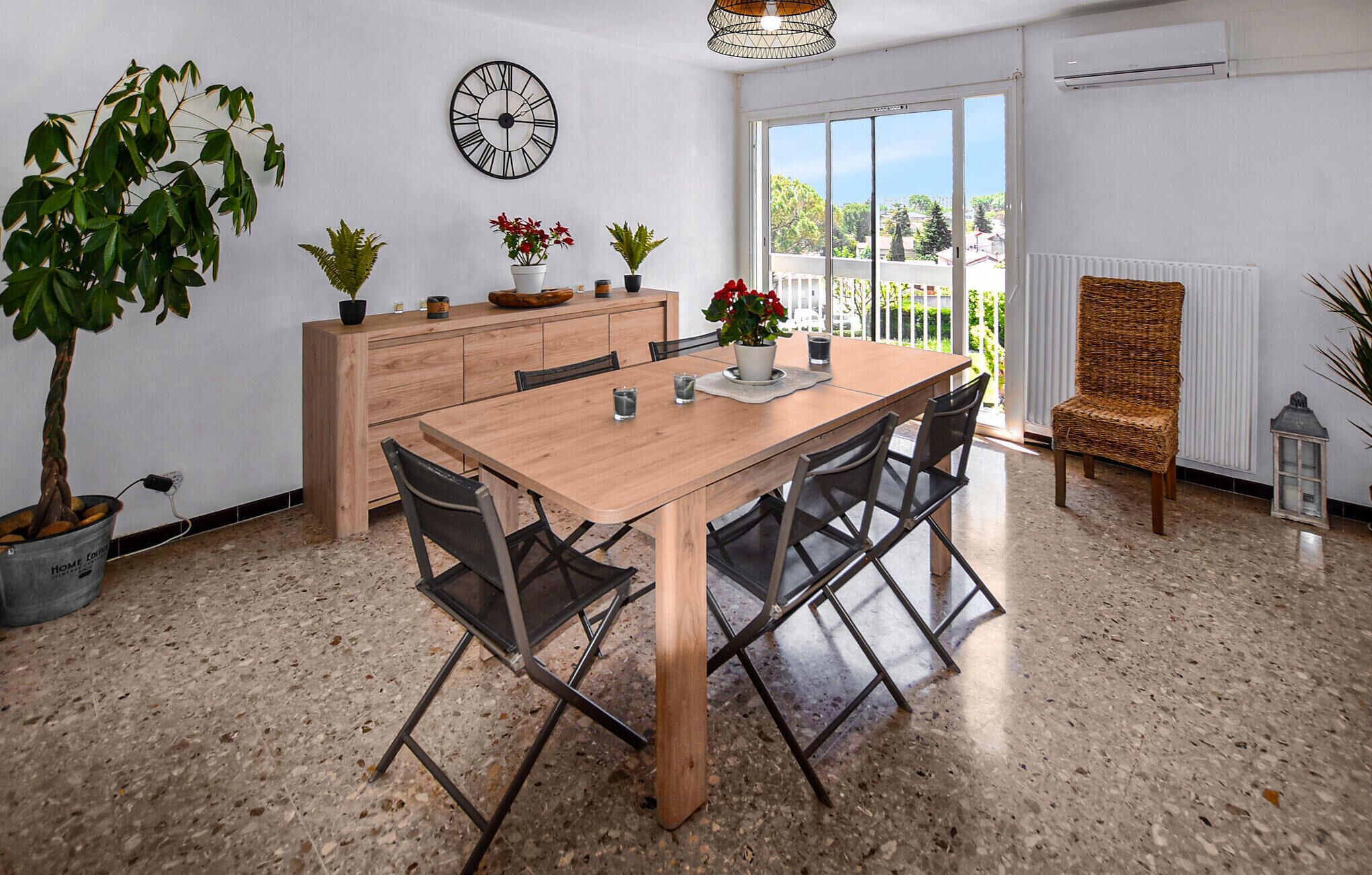 Een eethoek met een houten tafel, stoelen, potplanten en een wandklok.