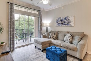 Ein Wohnzimmer mit einer Eckcouch, einem blauen Pouf, einer Stehlampe und einem großen Fenster mit Vorhängen.