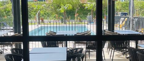 Un espacio de comedor al aire libre con mesas y sillas, una piscina al fondo y vistas a zonas verdes y un edificio en la calle.
