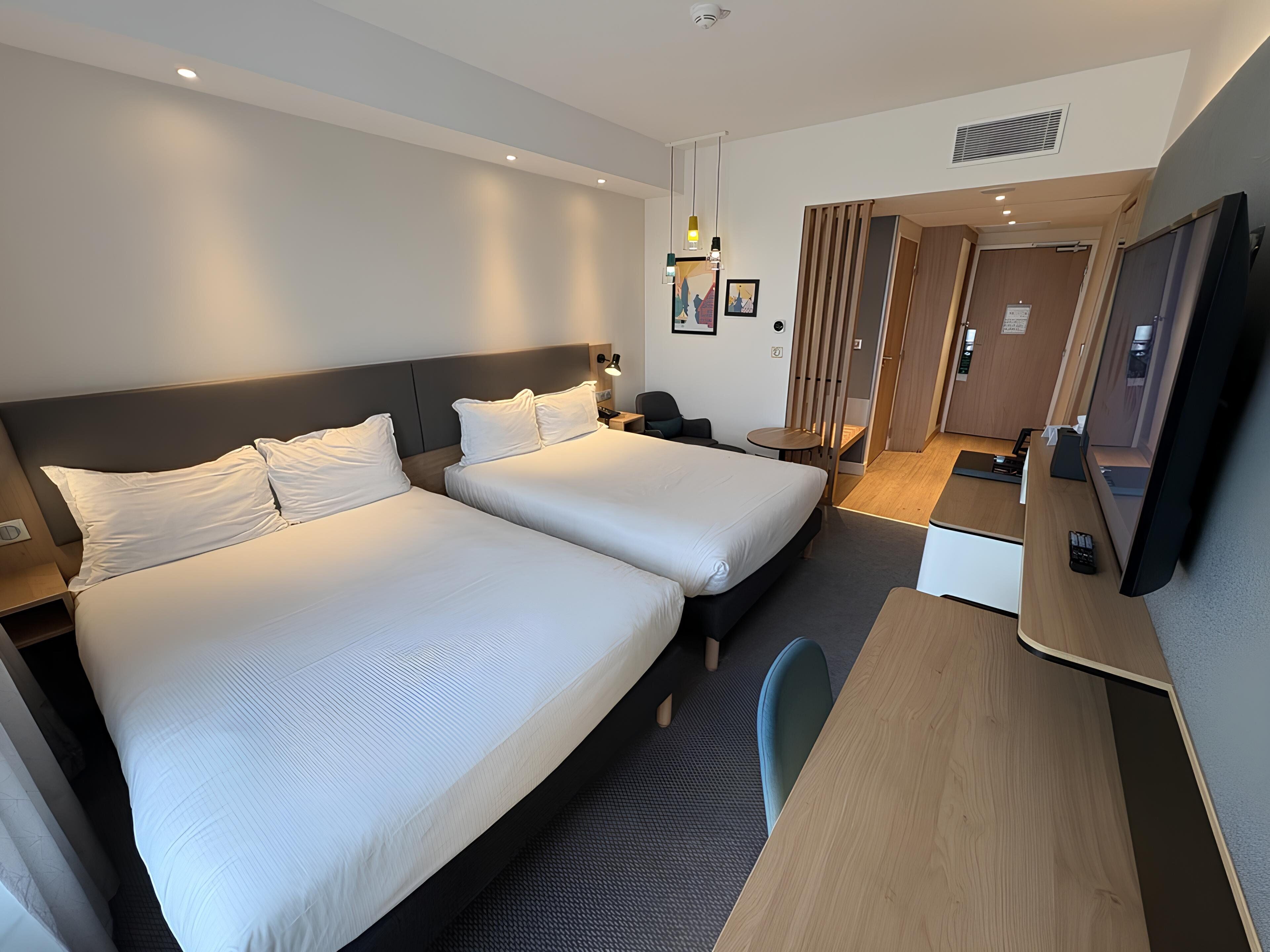 A hotel room with two beds, a desk, a chair, a TV, and a small table.
