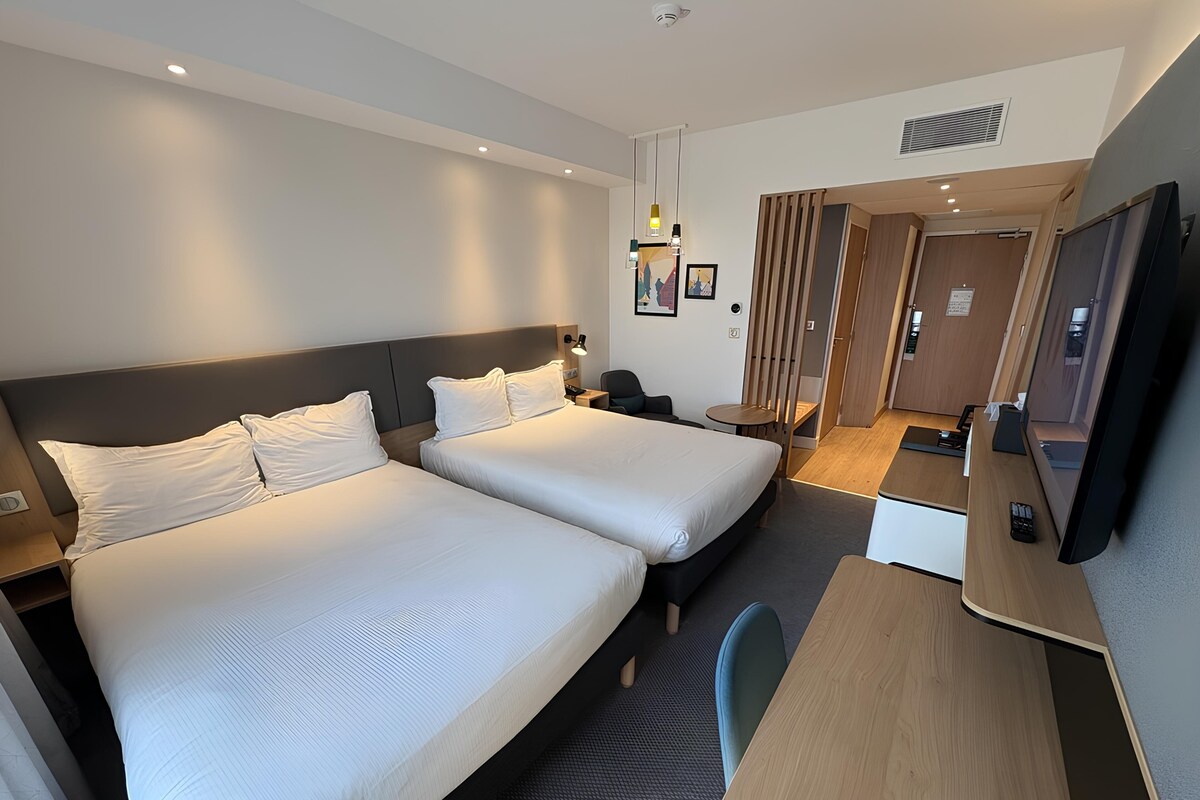 A hotel room with two beds, a desk, a chair, a TV, and a small table.