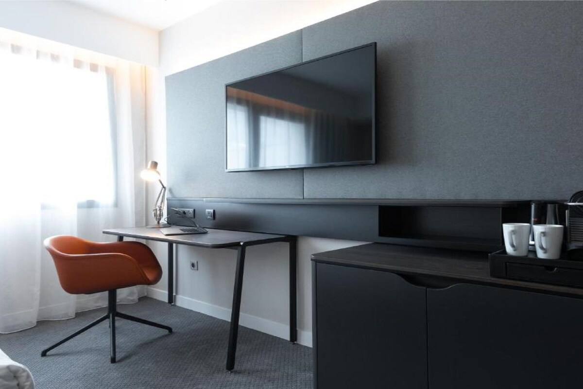 A modern hotel room with a desk, a comfortable chair, a flat-screen TV mounted on the wall, and a minibar.