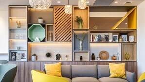 A modern living area with a sofa, yellow chairs, and a round wooden table. Shelves with decorative items and books are visible. - Holiday Inn Dijon Sud Longvic by IHG (Longvic)