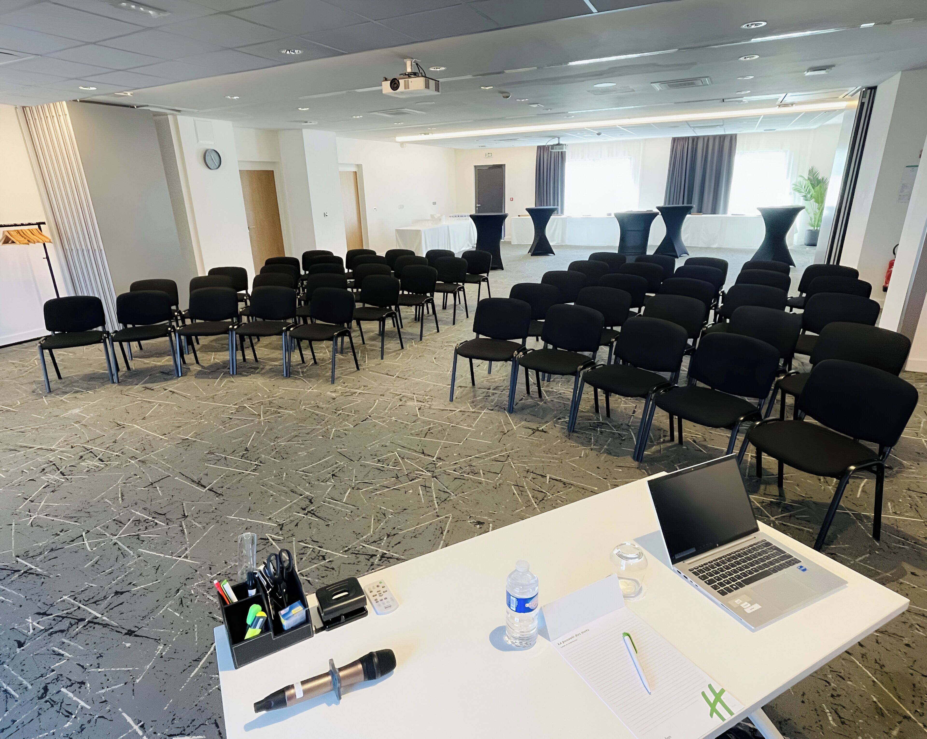 Une salle de conférence avec des rangées de chaises noires, une table blanche sur laquelle se trouve un ordinateur portable, un projecteur et une bouteille d’eau.