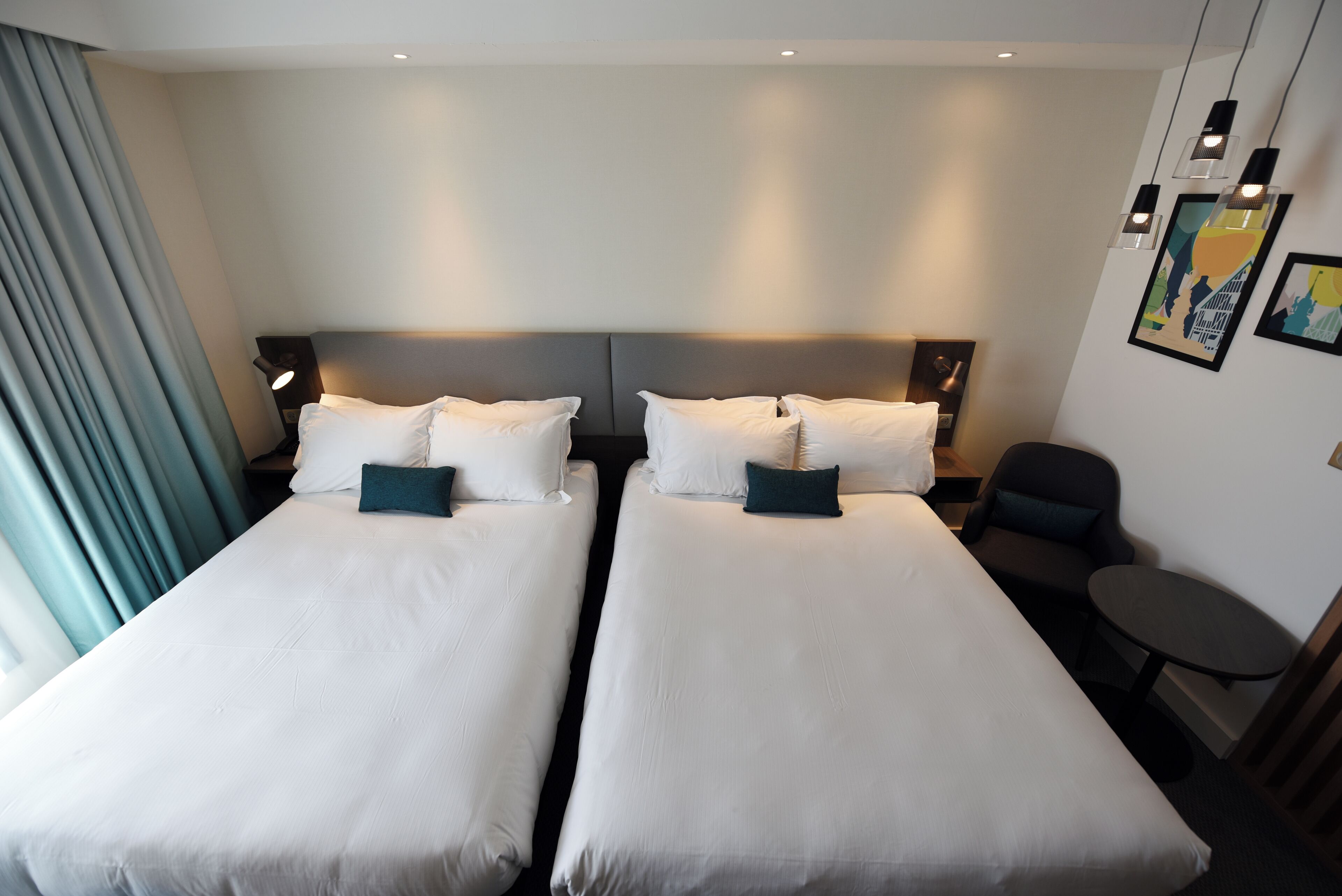 A hotel room with two single beds, a small table, a chair, and a nightstand with a lamp.