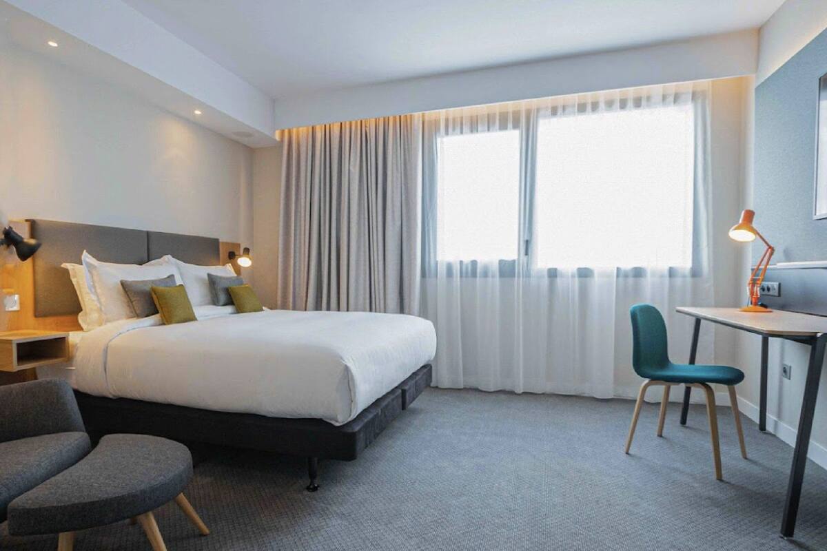 A hotel room with a bed, a desk, a chair, and a small table.