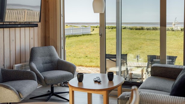 Une pièce moderne dotée d’une grande porte vitrée donnant sur un espace extérieur verdoyant, de deux fauteuils gris, d’une petite table ronde avec deux télécommandes et d’un luminaire suspendu blanc.