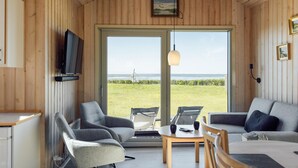 Un intérieur en bois avec un téléviseur, deux fauteuils gris, un canapé, une table et une grande porte-fenêtre coulissante donnant sur un espace verdoyant.