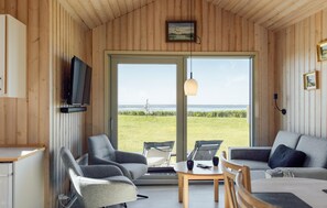 Un interior de madera con televisor, dos sillones grises, un sofá, una mesa y un amplio ventanal corredero que da a una zona verde.