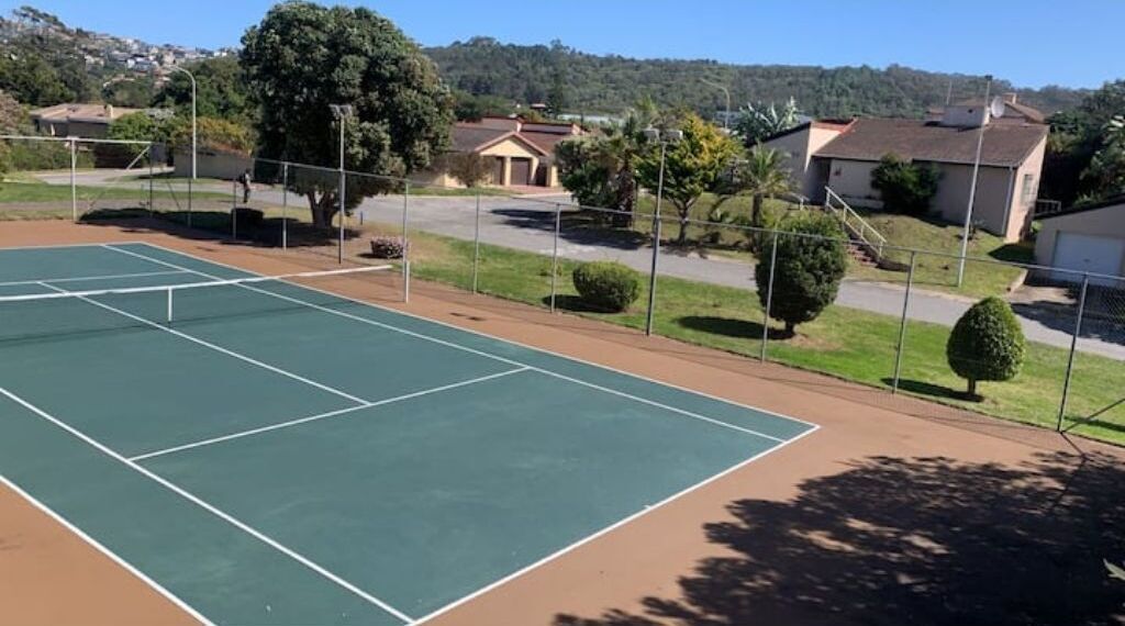A tennis court with a net, surrounded by a fence, and residential houses in the background.