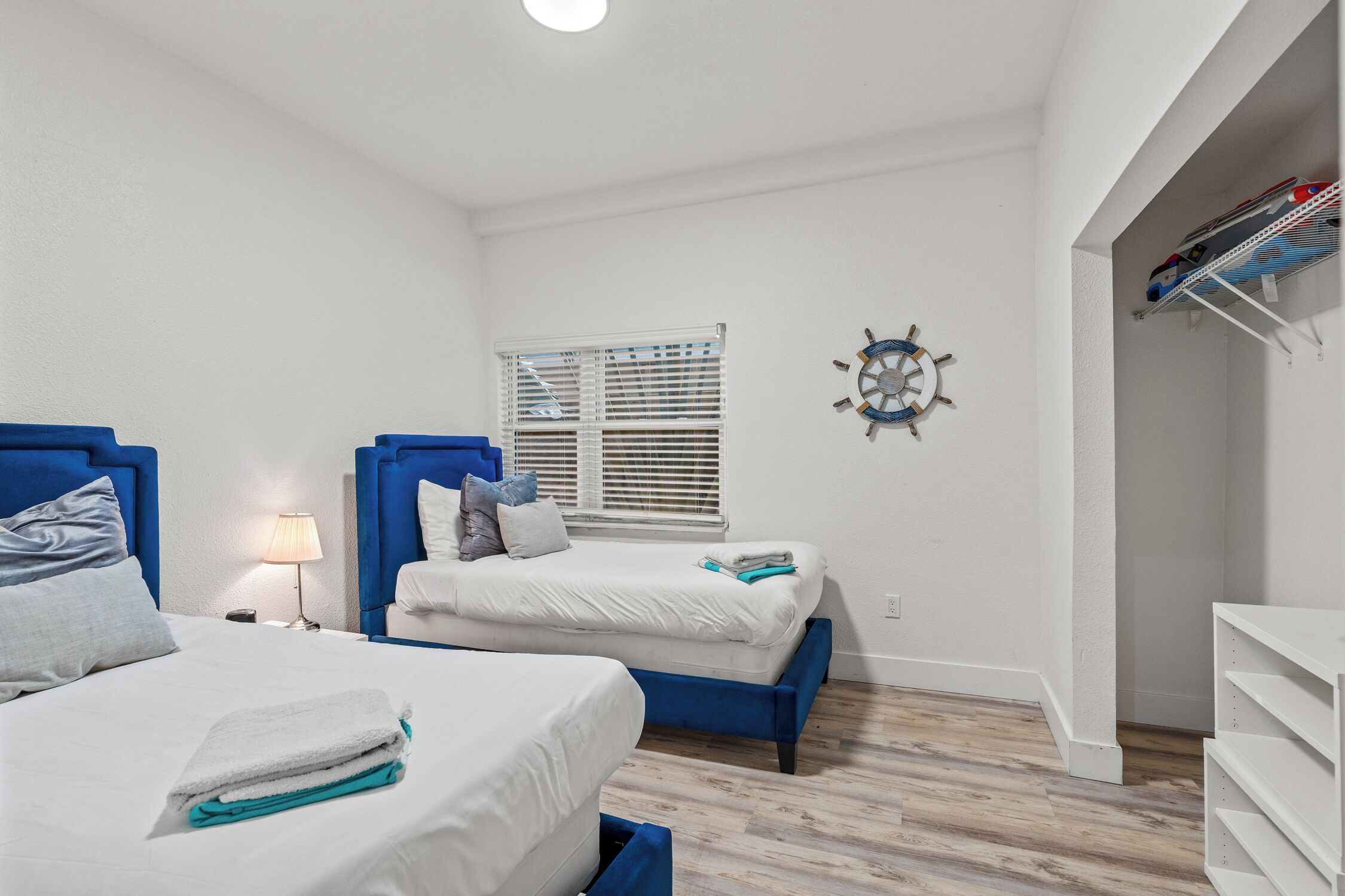 A hotel room with two beds, a window with blinds, a wall clock, and a shelf with books.