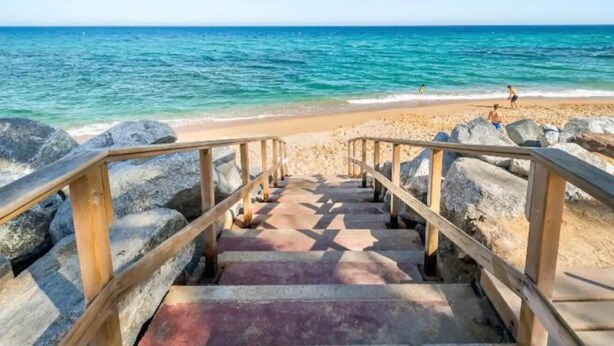 A wooden staircase leading to a beach with clear blue water and rocks. - Apartament Sant Mateu (Vilassar de Mar)