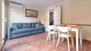 A room with a sofa, a dining table with chairs, a painting on the wall, and a door leading to another room. - Apartament Sant Mateu (Vilassar de Mar)