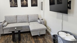 Ein Wohnzimmer mit einem grauen Ecksofa, einem flachbildfernseher an der Wand und drei gerahmten Bildern vom Eiffelturm.