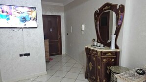 A room with a tiled floor, a wooden door, a TV mounted on the wall, a decorative mirror, and a small table with a vase. - Appartement (Tangier)