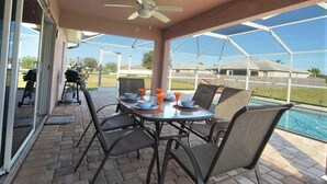 Une terrasse extérieure avec une table et des chaises, un ventilateur de plafond, et une vue sur une piscine et des maisons en arrière-plan.