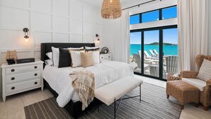 A bedroom with a large bed, a nightstand, a wicker chair, and a view of the ocean through large windows. - CIESTA SANDS- Beachfront Luxury, Top Beach, 4 bedrooms w private bath, Pool (Moore Hill)